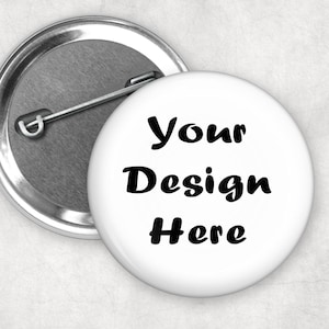 May include: A circular white pin-back button featuring the black text Your Design Here printed on the front. The metallic back of the button is visible, showing a silver-toned finish with a metal pin mechanism attached for fastening to clothing.