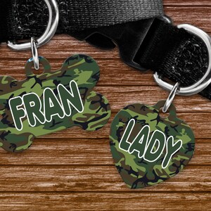 May include: Two pet ID tags with a camouflage pattern. One tag is bone-shaped and reads "FRAN" in white letters. The other tag is heart-shaped and reads "LADY" in white letters. Both tags are attached to a black dog collar.
