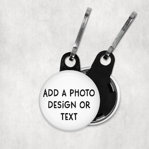 May include: White, round keychain tag with the text "ADD A PHOTO DESIGN OR TEXT" in black. The tag has a black backing and a silver-colored clip for attachment. The background is a textured, off-white color.