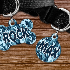 May include: Two dog tags with a blue camouflage pattern. One tag is bone-shaped and reads "ROCK" in white letters with a dark blue outline. The other tag is round and reads "MAX" in white letters with a dark blue outline.