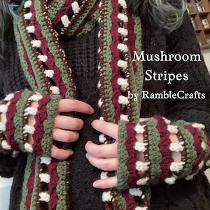 Puede incluir: Una bufanda y calentadores de muñeca a juego tejidos a crochet en rayas verdes, marrones, blancas y burdeos con un punto texturizado. La bufanda y los calentadores de muñeca se llaman "Mushroom Stripes" de RambleCrafts.