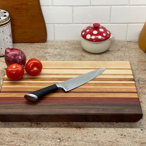 Puede incluir: Una tabla de cortar de madera con una variedad de tipos de madera, incluyendo nogal, arce y cerezo. La tabla tiene un acabado natural y mide aproximadamente 38 cm de largo y 25 cm de ancho. Un cuchillo de chef est&aacute; apoyado en la tabla, y hay dos tomates rojos y una cebolla roja en la tabla.