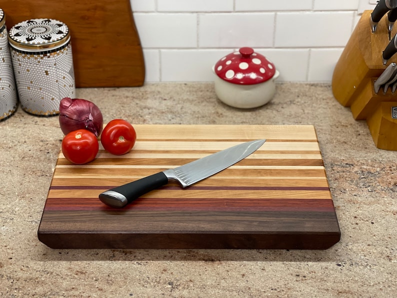 Puede incluir: Una tabla de cortar de madera con una variedad de tipos de madera, incluyendo nogal, arce y cerezo. La tabla tiene un acabado natural y mide aproximadamente 38 cm de largo y 25 cm de ancho. Un cuchillo de chef est&aacute; apoyado en la tabla, y hay dos tomates rojos y una cebolla roja en la tabla.