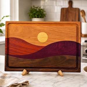 May include: A rectangular wooden cutting board featuring an inlaid landscape design. The board displays a yellow sun against a light wood sky, with rolling hills in shades of reddish-orange, deep purple, and dark brown wood grain.