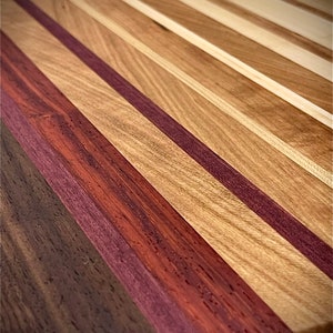 Puede incluir: Una tabla de cortar de madera con una variedad de tipos de madera, incluyendo nogal, arce y palo de rosa. La tabla est&aacute; hecha de tiras de madera alternas, creando un patr&oacute;n visualmente atractivo.