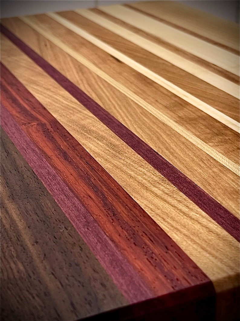 Puede incluir: Una tabla de cortar de madera con una variedad de tipos de madera, incluyendo nogal, arce y palo de rosa. La tabla est&aacute; hecha de tiras de madera alternas, creando un patr&oacute;n visualmente atractivo.