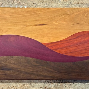 May include: A rectangular wooden cutting board featuring a multi-colored inlay design. The board displays horizontal, wavy sections of light tan, deep purple, reddish-orange, and dark brown wood grain, creating a layered, landscape-like pattern.