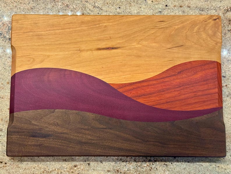 May include: A rectangular wooden cutting board featuring a multi-colored inlay design. The board displays horizontal, wavy sections of light tan, deep purple, reddish-orange, and dark brown wood grain, creating a layered, landscape-like pattern.