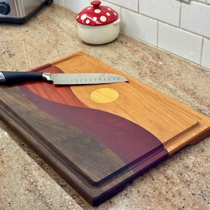 May include: A rectangular wooden cutting board featuring a multi-colored landscape design with wavy layers of dark brown, deep purple, and light tan wood, accented by a circular yellow inlay. A stainless steel kitchen knife rests on top.