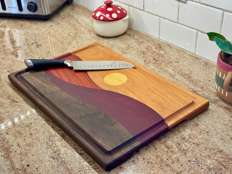 May include: A rectangular wooden cutting board featuring a multi-colored landscape design with wavy layers of dark brown, deep purple, and light tan wood, accented by a circular yellow inlay. A stainless steel kitchen knife rests on top.