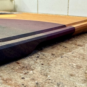 May include: A rectangular wooden cutting board featuring a two-tone design with a light tan wood section and a dark purple wood section. The board includes a carved juice groove along the perimeter and a recessed handle cutout on the side edge.
