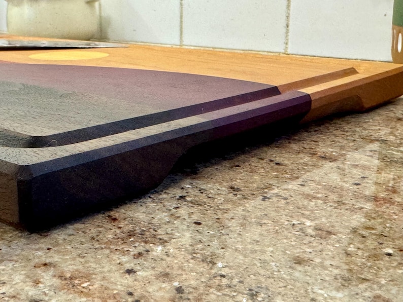 May include: A rectangular wooden cutting board featuring a two-tone design with a light tan wood section and a dark purple wood section. The board includes a carved juice groove along the perimeter and a recessed handle cutout on the side edge.