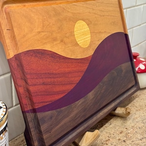 May include: A rectangular wooden cutting board featuring an inlaid landscape design. The board displays a light-colored wood sky with a circular sun inlay, above rolling hills crafted from deep red, purple, and dark brown wood segments.