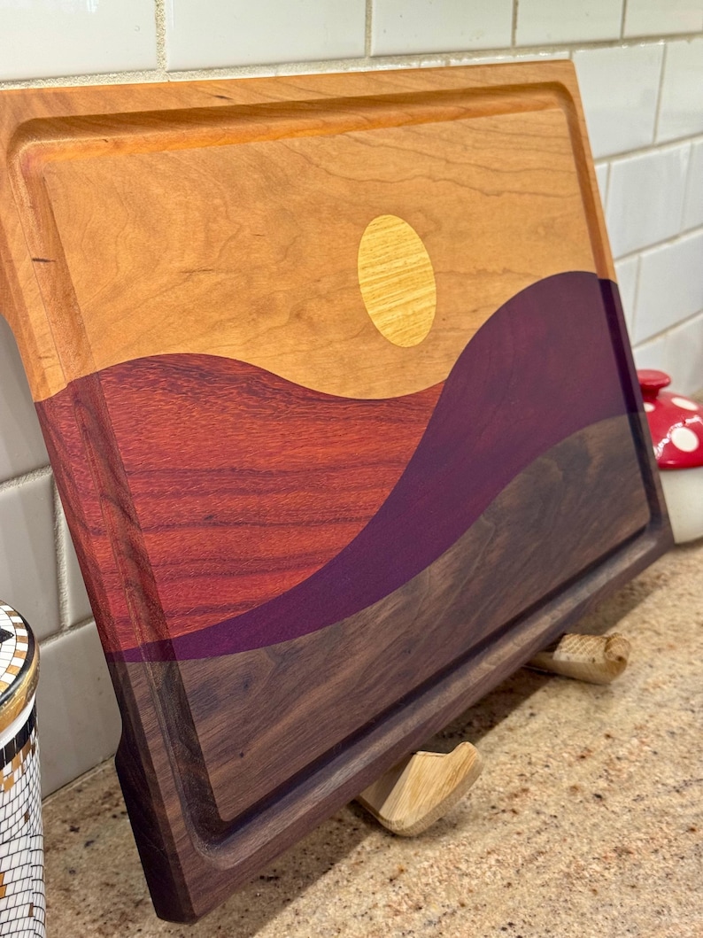May include: A rectangular wooden cutting board featuring an inlaid landscape design. The board displays a light-colored wood sky with a circular sun inlay, above rolling hills crafted from deep red, purple, and dark brown wood segments.