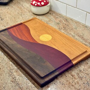 May include: A rectangular wooden cutting board featuring a multi-colored inlay design of rolling hills and a sun. The board is constructed from contrasting dark brown, deep purple, reddish-brown, and light tan wood segments with a juice groove.