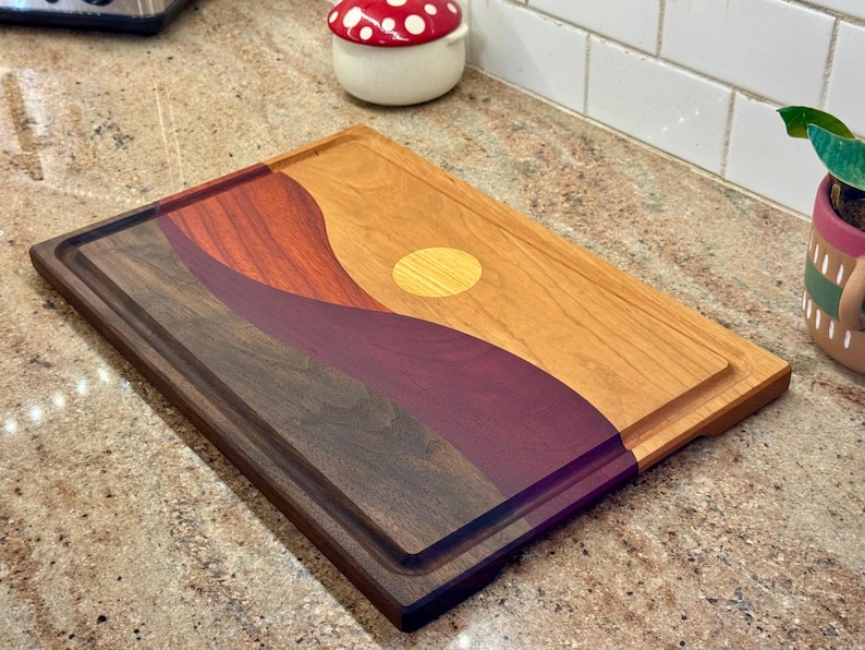 May include: A rectangular wooden cutting board featuring a multi-colored inlay design of rolling hills and a sun. The board is constructed from contrasting dark brown, deep purple, reddish-brown, and light tan wood segments with a juice groove.