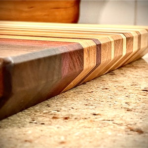Puede incluir: Primer plano de una tabla de cortar de madera con un patr&oacute;n de chevron hecho de diferentes tipos de madera. La tabla tiene un esquema de color marr&oacute;n oscuro, marr&oacute;n rojizo y marr&oacute;n claro.