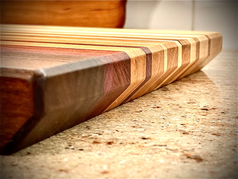 Puede incluir: Primer plano de una tabla de cortar de madera con un patr&oacute;n de chevron hecho de diferentes tipos de madera. La tabla tiene un esquema de color marr&oacute;n oscuro, marr&oacute;n rojizo y marr&oacute;n claro.