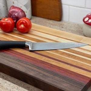 Puede incluir: Una tabla de cortar de madera grande con una variedad de tipos de madera, incluyendo nogal, arce y cerezo. La tabla tiene un acabado natural y est&aacute; dise&ntilde;ada para su uso en la cocina. Un cuchillo plateado est&aacute; apoyado en la tabla.