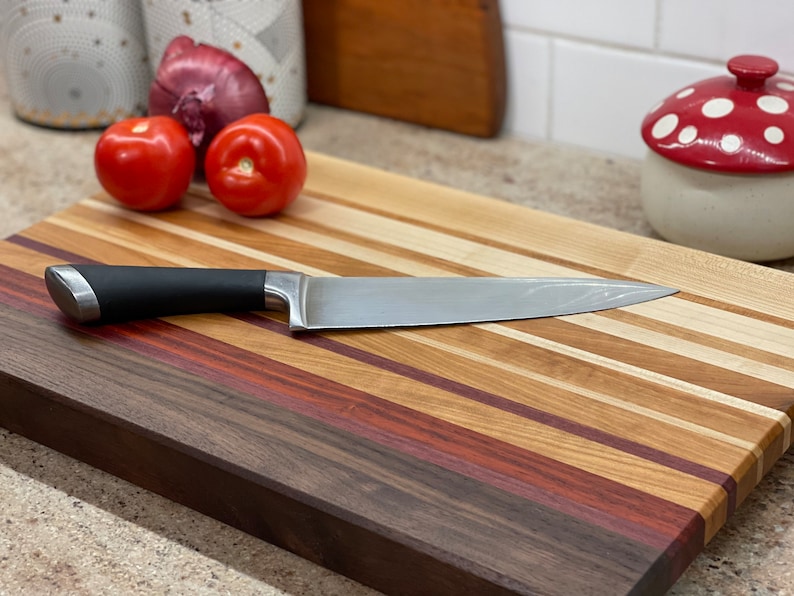 Puede incluir: Una tabla de cortar de madera grande con una variedad de tipos de madera, incluyendo nogal, arce y cerezo. La tabla tiene un acabado natural y est&aacute; dise&ntilde;ada para su uso en la cocina. Un cuchillo plateado est&aacute; apoyado en la tabla.