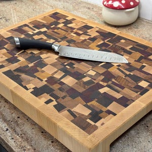 May include: A rectangular end-grain cutting board with a mosaic pattern of various wood tones, including light brown, dark brown, and purple. A stainless steel chef's knife with a black handle rests on the board. The board is approximately 18 inches long.