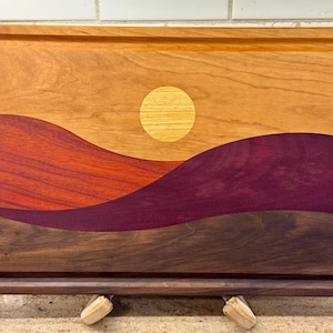 May include: A rectangular wooden cutting board featuring an inlaid landscape design. The board displays a light wood sky with a circular yellow sun, layered over rolling hills in shades of reddish-orange, deep purple, and dark brown wood grain.