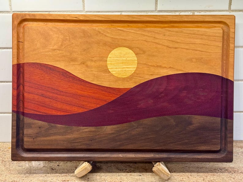 May include: A rectangular wooden cutting board featuring an inlaid landscape design. The board displays a light wood sky with a circular yellow sun, layered over rolling hills in shades of reddish-orange, deep purple, and dark brown wood grain.