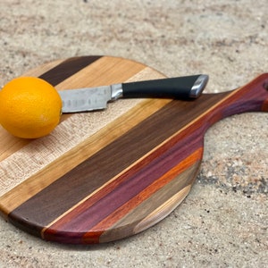 May include: A round wooden cutting board with a handle, featuring a variety of wood tones in diagonal stripes. A small knife with a black handle rests on the board, next to a whole orange. The board is approximately 12 inches in diameter.