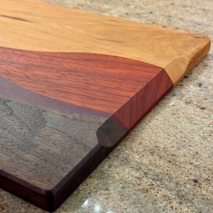 May include: A rectangular wooden cutting board featuring a multi-tonal design with distinct sections of light tan, reddish-brown, and dark brown wood grain. The board has smooth, beveled edges and a polished finish, showcasing the natural wood texture.