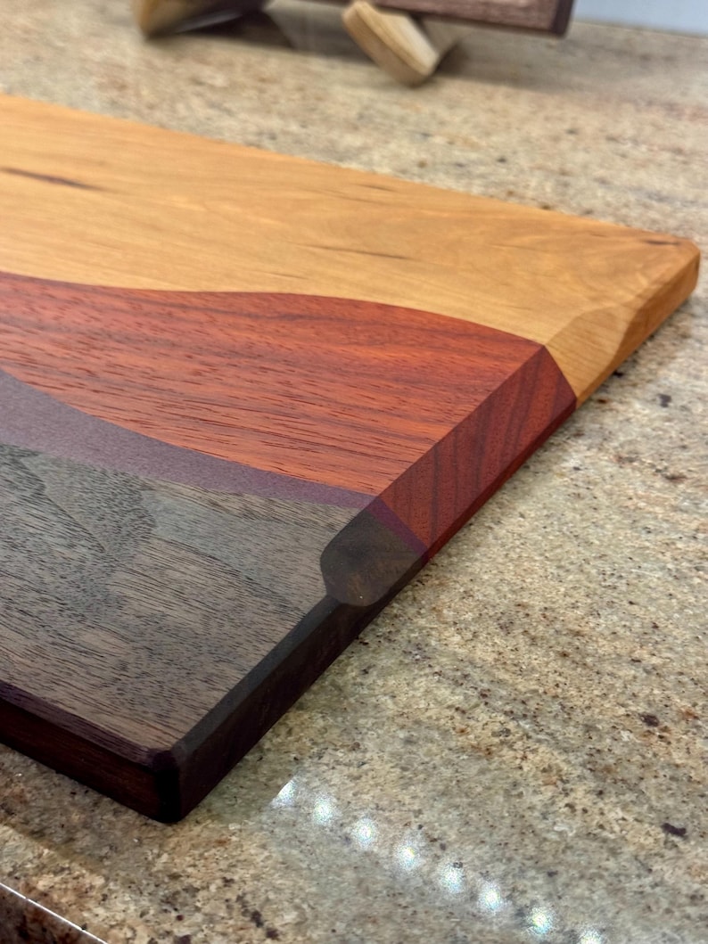May include: A rectangular wooden cutting board featuring a multi-tonal design with distinct sections of light tan, reddish-brown, and dark brown wood grain. The board has smooth, beveled edges and a polished finish, showcasing the natural wood texture.