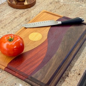 May include: A rectangular wooden cutting board featuring a multi-toned, wavy inlay design in shades of light tan, deep red, and dark brown. A single red tomato and a stainless steel kitchen knife with a black handle rest on the board surface.
