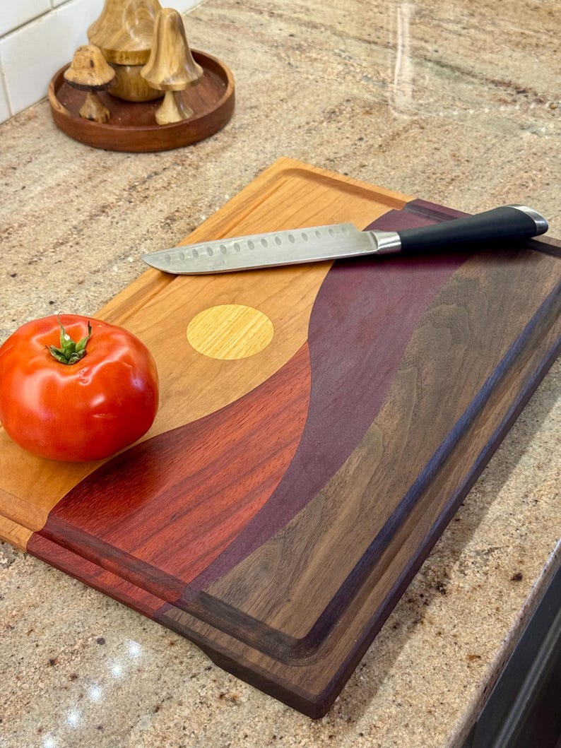 May include: A rectangular wooden cutting board featuring a multi-toned, wavy inlay design in shades of light tan, deep red, and dark brown. A single red tomato and a stainless steel kitchen knife with a black handle rest on the board surface.