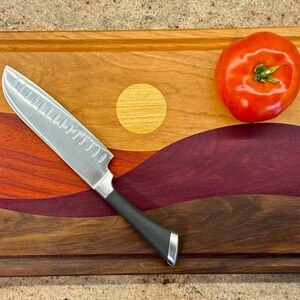 May include: A rectangular wooden cutting board featuring a multi-colored inlay design of a landscape with a yellow sun, red and purple hills, and a dark brown base. A silver santoku knife with a black handle and a red tomato rest on the board.