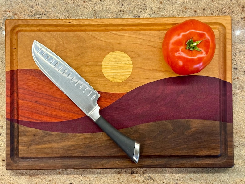 May include: A rectangular wooden cutting board featuring a multi-colored inlay design of a landscape with a yellow sun, red and purple hills, and a dark brown base. A silver santoku knife with a black handle and a red tomato rest on the board.