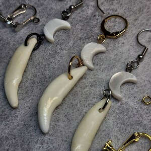 May include: Three pairs of earrings with a white tooth and crescent moon design. The earrings are made with gold and silver metal.