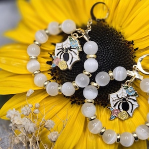 May include: Two white beaded bracelets with gold accents and a black spider charm with colorful flowers. The bracelets are on a yellow sunflower.