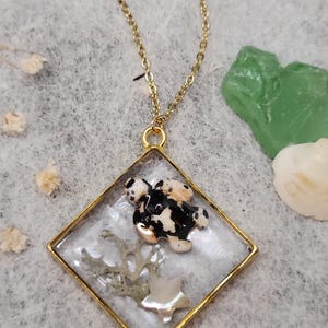 May include: A gold-toned necklace featuring a clear, square pendant. Inside the pendant is a black and white speckled turtle-shaped charm, a piece of dried plant, and a mother-of-pearl star. The necklace is on a light gray surface.