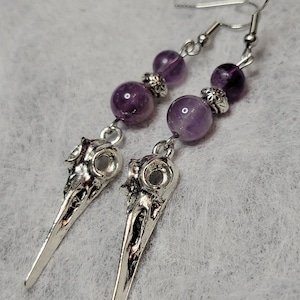 May include: A pair of silver skull earrings with amethyst beads. The skulls are detailed and have a pointed shape. The amethyst beads are a deep purple colour.