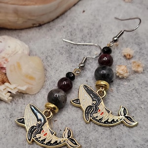 May include: A pair of whale earrings with black and white enamel whale charms, gold-toned metal, and dark grey, burgundy, and black beads.