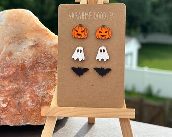 May include: A wooden easel displays a card with Halloween-themed earrings. The earrings include orange pumpkins, white ghosts, and black bats. The card has the text "SARAHME DOODLES" at the top. The easel is on a surface with a blurred background.