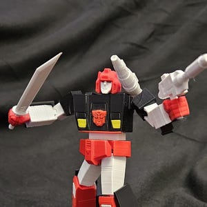 May include: A 3D-printed action figure in a dynamic pose, featuring a red helmet and accents, black torso, and white and red limbs. It holds a white sword in one hand and a white gun in the other. The figure has a detailed design and is set against a dark background.