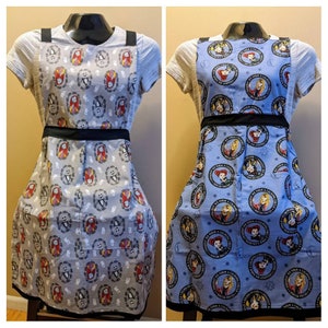 May include: Two aprons on mannequins. One is light gray with a pattern of characters from The Nightmare Before Christmas. The other is light blue with a pattern of characters from Hocus Pocus. Both aprons have black straps and waistbands.