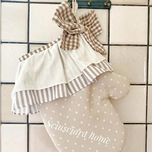 Könnte beinhalten: Ein beiger Ofenhandschuh mit weißen Punkten und dem Text "Sciusciara home". Er hat eine gerüschte Bordüre mit Streifen und eine braune Vichy-Schleife. Der Handschuh hängt an einer gefliesten Wand.