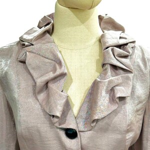 K&ouml;nnte beinhalten: Ein helllila Blazer mit R&uuml;schenkragen und einem einzelnen schwarzen Knopf. Der Stoff hat einen dezenten Glanz, und die Jacke wird auf einer Schaufensterpuppe pr&auml;sentiert. Das Design ist klassisch und elegant.
