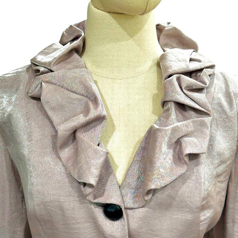 K&ouml;nnte beinhalten: Ein helllila Blazer mit R&uuml;schenkragen und einem einzelnen schwarzen Knopf. Der Stoff hat einen dezenten Glanz, und die Jacke wird auf einer Schaufensterpuppe pr&auml;sentiert. Das Design ist klassisch und elegant.