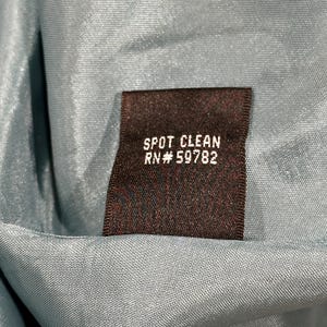 Op de afbeelding: Close-up van een donkerbruin label met de woorden "SPOT CLEAN" en "RN# 59782" in het wit gedrukt. Het label staat tegen een lichtblauwe stofachtergrond.