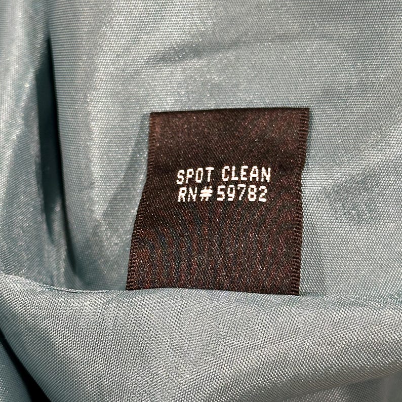 Op de afbeelding: Close-up van een donkerbruin label met de woorden "SPOT CLEAN" en "RN# 59782" in het wit gedrukt. Het label staat tegen een lichtblauwe stofachtergrond.