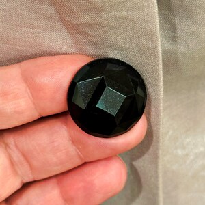 K&ouml;nnte beinhalten: Ein gro&szlig;er, facettierter schwarzer Knopf mit geometrischem Design. Der Knopf ist rund und hat eine gl&auml;nzende Oberfl&auml;che. Der Knopf wird vor einem hellbraunen Stoff gehalten.