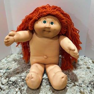May include: A soft-sculpture doll with a round face, dimpled cheeks, and a small belly button. The doll features long, textured orange yarn hair, green eyes, and a light tan fabric body. The arms are extended outward, and the legs are positioned in a seated pose.