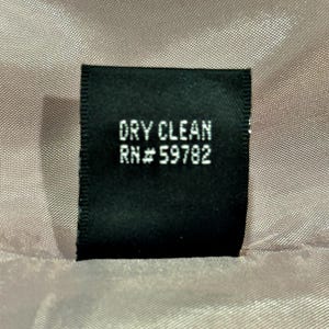 K&ouml;nnte beinhalten: Ein schwarzes Etikett mit dem wei&szlig;en Aufdruck "DRY CLEAN" und "RN# 59782". Das Etikett befindet sich vor einem strukturierten, hellrosa Stoff.